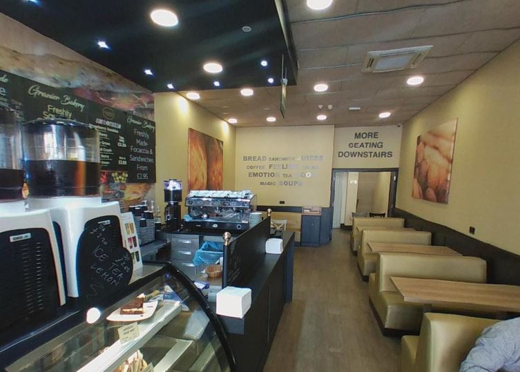 The interior looking down the length of the room: an espresso machine and glass display cabinet on the left, upholstered booth seating with wooden tables on the right, and a wall sign reading More Seating Downstairs.