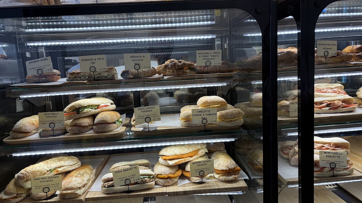 Three lit shelves of a chilled cabinet holding filled focaccias, baguettes, bagels, rolls and savoury croissants, each on a wooden board with a printed name card in front of it.